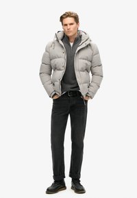 Chaqueta acolchada gris con capucha, diseño acolchado, llevada sobre un suéter gris oscuro con cremallera, combinada con jeans oscuros y botas negras con cordones.
