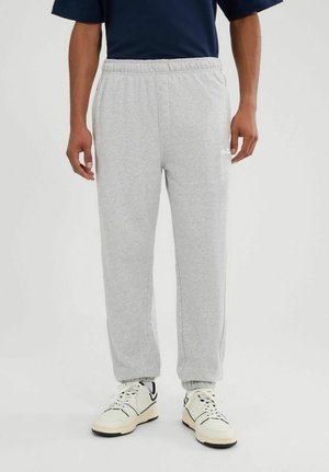 Hombre con pantalones deportivos gris claro con cintura y puños elásticos, zapatillas blancas con detalles en negro, de pie frente a un fondo liso.