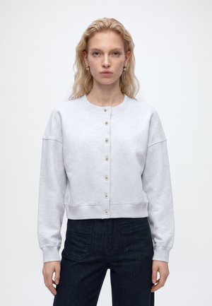 Blondynka o falowanych włosach ma na sobie jasnoszarą sweterkę na guziki i ciemne dżinsy, stoi na tle jednolitego białego tła.