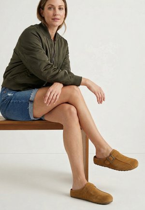 Kvinde iført grøn bomberjakke, denimshorts og brune slip-on træsko, siddende på en træbænk mod en ensfarvet baggrund.