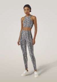 Vrouw staat, gekleed in bijpassende zwart-witte slangenprint crop top en leggings met witte sneakers, neutrale achtergrond.