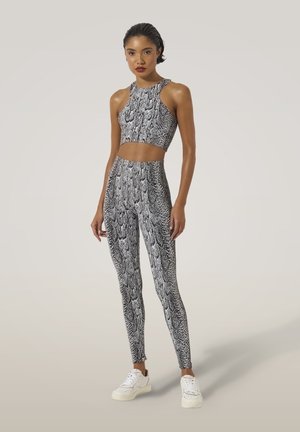 Woman standing, wearing matching black and white snake-print crop top and leggings with white sneakers, neutral background.