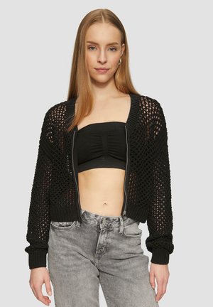 Zwarte cropped cardigan met een opengehaakt ontwerp, voorzien van een ritssluiting aan de voorkant en geribde manchetten. Gemaakt over een zwarte aansluitende top en een grijze jeans.