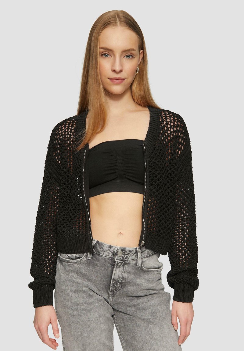 Schwarze, kurz geschnittene Strickjacke mit Offene-Strickdesign, ausgestattet mit einem Reißverschluss an der Vorderseite und gerippten Bündchen. Getragen über einem schwarzen, figurbetonten Oberteil und grauen Jeans.