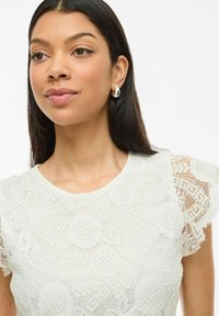 Hvid blonde top med blomsterprint, bølgede ærmer og rund halsudskæring. Model bærer sølvøreringe og har lige sort hår.