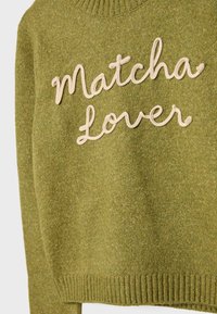 Grøn strikket sweater med ribbet halsudskæring og manchetter. "Matcha Lover" er broderet med beige tråd hen over fronten.