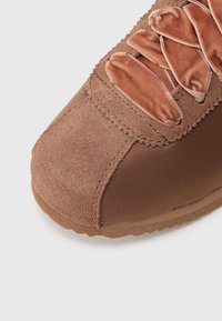 Brązowy but sportowy z zamszową cholewką i teksturowaną tkaniną, z płaskimi sznurowadłami z tkaniny i gumową podeszwą, o zaokrąglonym kształcie noska.