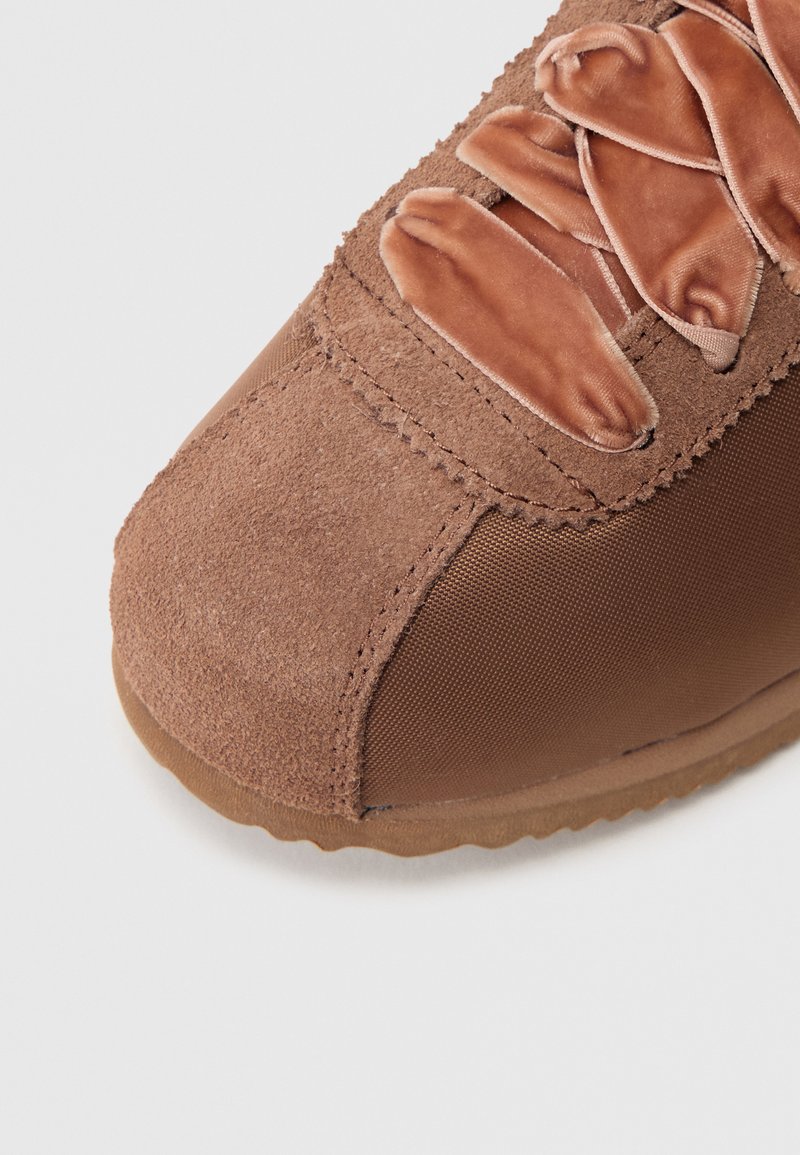 Brown athletic shoe featuring a suede upper and textured fabric, with flat fabric laces and a rubber outsole, showcasing a rounded toe design.