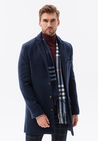 Mörkblå ullblandad kappa med slagkrage, framfickor och sex knappar. Matchad med en mönstrad grå och blå scarf.
