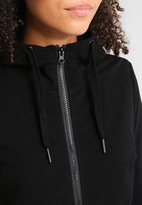 Sudadera con cremallera negra que cuenta con un cuello alto, cordones y una textura suave. La cremallera es de color plateado con un acabado mate.