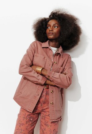 Femme aux cheveux bouclés et volumineux, portant une veste à boutons rose poudré et un pantalon à motifs floraux, les bras croisés, regardant sur le côté.