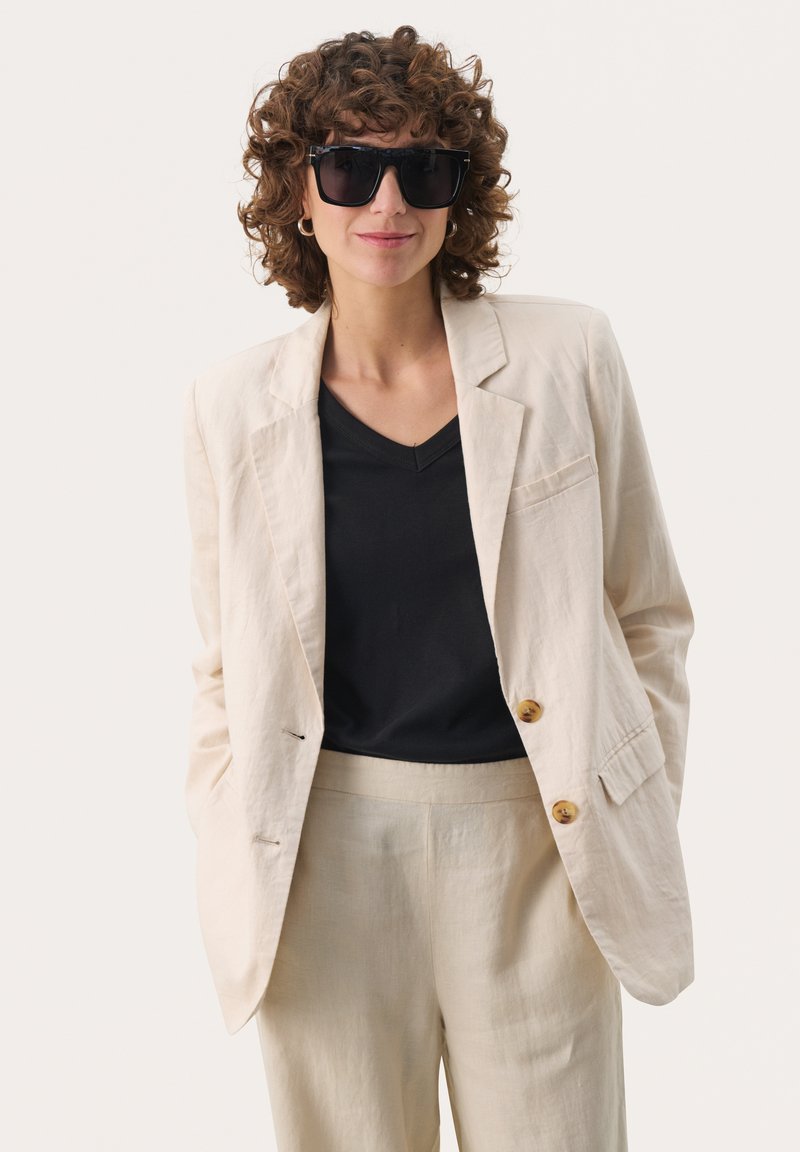 Beige linneblazer med två knappar, i kombination med en svart V-ringad topp och ljusbeiga byxor. Modell med stora svarta solglasögon.