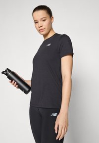 T-shirt de sport noir à manches courtes avec un petit logo réfléchissant, associé à un legging noir présentant un logo similaire, tenu avec une bouteille d'eau noire.