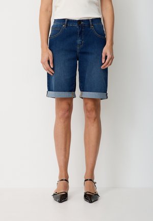 Frau steht und trägt dunkelblaue, hochgekrempelte Jeans-Shorts, ein weißes Oberteil und schwarze Schuhe mit spitzer Spitze und Knöchelriemen vor weißem Hintergrund.