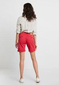 Pantalones cortos de algodón rojos con dobladillos, que presentan bolsillos traseros y una pequeña etiqueta de la marca. Combinados con una camisa de lino de color claro y zapatillas blancas.