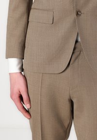 Abito marrone chiaro, tessuto testurizzato, con giacca a un bottone, revers a lancia e vestibilità su misura; polsino della camicia bianca visibile sotto la manica.