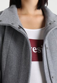 Giacca in misto lana grigia con collo a camicia, bottoni a pressione e una texture liscia, indossata sopra una camicia bianca con lettering bordeaux.