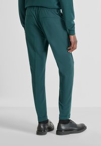 Zelene hlače s elastičnim pojasom, suženim krojem i manžetama, uparene s crnim cipelama. Sadrži dvije stražnje džepove.