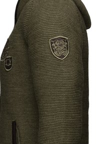 Veste en tricot à capuche de couleur vert olive, présentant un motif texturé, plusieurs badges et deux poches latérales zippées.