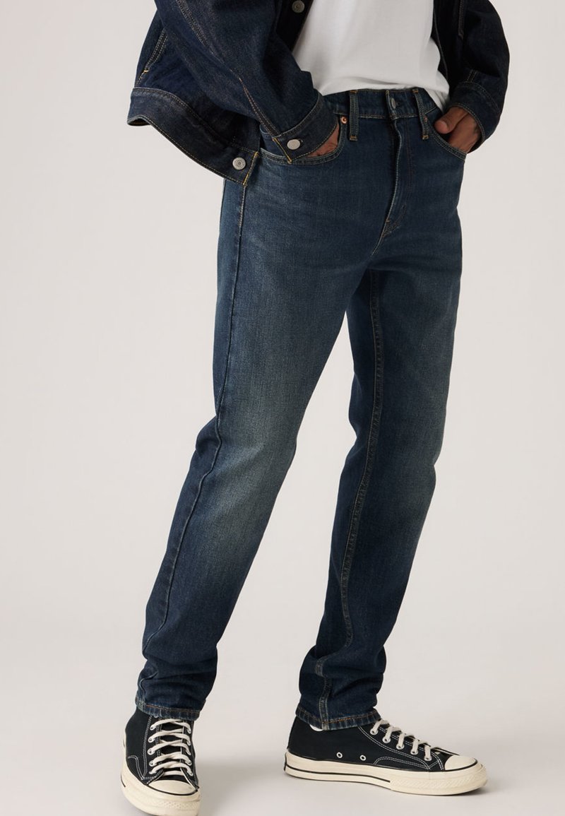 Jean en denim bleu foncé à coupe droite, avec un design à cinq poches et des coutures contrastantes jaunes. Porté avec des baskets noires.