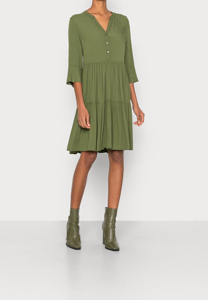 Femme portant une robe olive longueur genoux avec des manches trois-quarts et des bottines vertes à talons assorties, debout sur un fond uni.