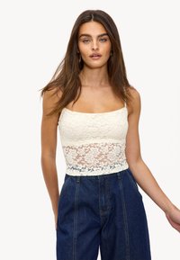 Top corto de encaje en color blanco roto con un patrón floral y tirantes finos, combinado con pantalones vaqueros de cintura alta en azul oscuro.