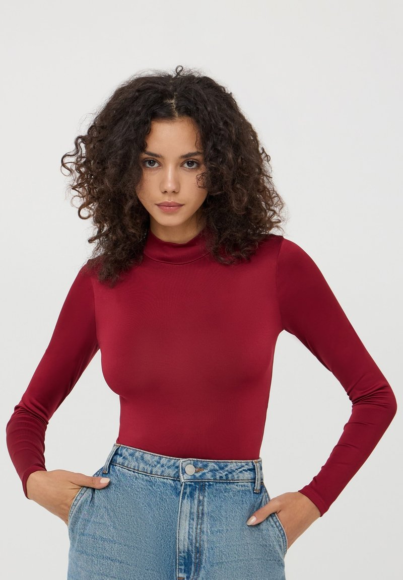 Maglia rossa a maniche lunghe con collo a imbuto in tessuto elasticizzato, con un look aderente. Abbinata a jeans in denim chiaro a vita alta.