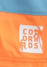 Färggt, lätt tyg med en orange botten och en blå överdel. Vit logga med palmträdaccentuerar centrerad på tyget.