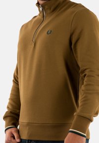 Sudadera marrón con cremallera y cuello alto, con puños y dobladillo acanalados, que presenta un pequeño logotipo bordado en azul oscuro.