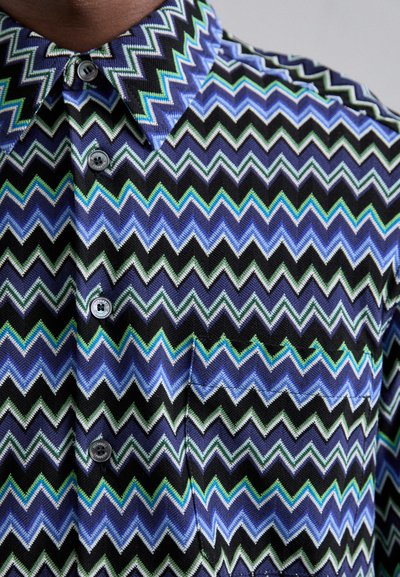 Κοντομάνικη πουκάμισο με σχέδιο ζιγκ-ζαγκ σε μοβ, μπλε, πράσινο και μαύρο χρώμα. Διαθέτει γιακά, δύο κουμπιά και τσέπη μπροστά.