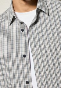 Geruit overhemd in beige en marineblauw, met een kraag en donkere knopen. Draag het over een witte basislaag.