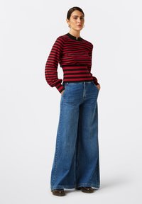 Gestreifter rot-schwarzer Strickpullover mit hohem Kragen, kombiniert mit weiten blauen Denim-Jeans. Entspannte Passform und schwingende Ärmel.