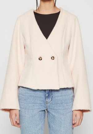 Frau trägt eine hellrosa, flauschige Doppelknopf-Cropped-Jacke über einem schwarzen Top und blauen Jeans, stehend vor einem einfarbigen Hintergrund.