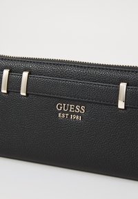 Black synthetic leather wallet featuring a textured finish, gold-tone hardware accents, and embossed logo with "EST 1981" below.