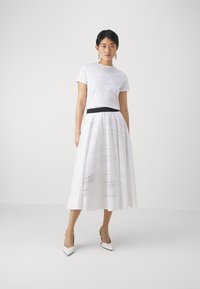 Tricou alb cu text imprimat, asociat cu o fustă midi albă brodată, lungă, cu modele decupate și un tiv negru; tocuri albe.