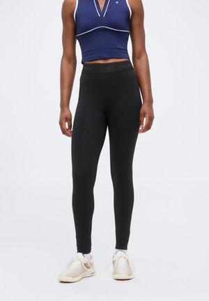 Oseba, ki nosi črne pajkice z visokim pasom, temno moder brezrokavnik z belim trakom in svetle tekaške copate, stoji pred enobarvnim ozadjem.