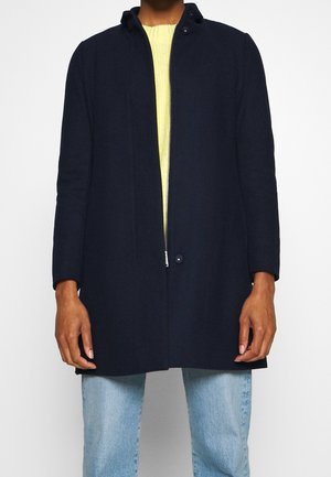 Personne portant un manteau zippé bleu marine par-dessus un pull jaune et un jean bleu clair sur un fond blanc uni.