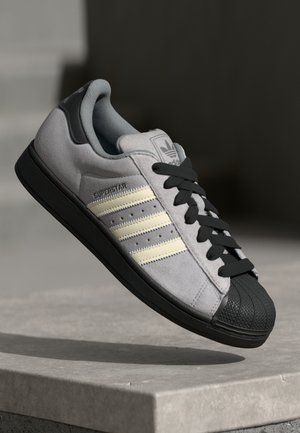 Gray suede sneaker with black rubber sole and toe guard, featuring three cream stripes and black laces, labeled "SUPERSTAR" on the side.
