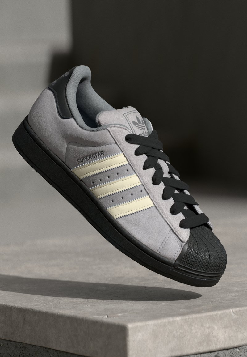 Šedé semišové sneakers s čiernou gumenou podrážkou a ochranou prstov, s tromi smotanovými pruhmi a čiernymi šnúrkami, označené ako "SUPERSTAR" na boku.