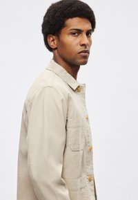 Beżowa zapinana na guziki kurtka z kołnierzem, pojedynczą kieszenią na piersi i luźnym krojem. Wykonana z lekkiego materiału z widocznymi przeszyciami.