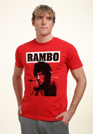 Rød bomulds t-shirt med et sort-hvidt grafik af en muskuløs karakters ansigt, mærket "RAMBO" med fed skrift i sort ovenfor.