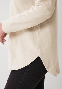 Beige Langarmshirt aus weichem Stoff, mit abgerundetem Saum und seitlichen Schlitzen. Kombiniert mit dunklen Jeans, die den Farbkontrast betonen.