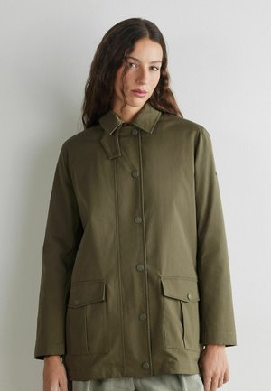 Frau trägt eine lange olivgrüne Jacke mit Knöpfen und Klapptaschen, steht vor einer schlichten hellgrauen Wand.