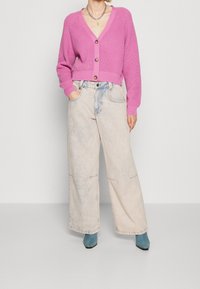 Fuksjowy sweter na drutach z dekoltem w serek, zapinany na trzy guziki. W zestawieniu z jasnymi, postrzępionymi, szerokimi jeansami i turkusowymi botkami za kostkę.