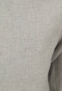 Graüer Stoff mit strukturierter Webart, ausgestattet mit einem gestickten "M©G"-Logo in heller Fadenfarbe. Nahaufnahme, die Material- und Nahtdetails hervorhebt.