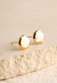 Goldene Ohrstecker mit abgerundeter, unregelmäßiger Form und glänzender Oberfläche, die auf einem strukturierten, hellfarbigen Steinuntergrund liegen.