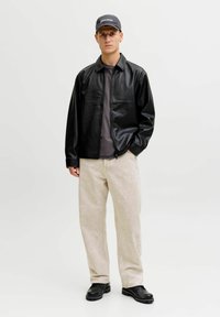 Schwarze Jacke aus Kunstleder, graues Hemd, hellbeige Hose und schwarze Stiefel. Das Model steht vor einem einfarbigen Hintergrund.