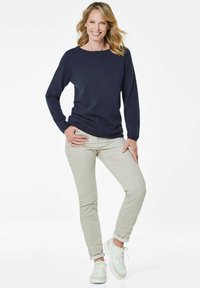 Dunkelblauer Pullover mit Rundhalsausschnitt, hellbeige Slim-Fit-Hosen und weißen Sneakers. Das Model posiert mit einer Hand auf der Hüfte und steht auf einem Bein.