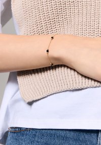 Złota bransoletka z czarnymi koralikami, o delikatnym wzorze, noszona na nadgarstku na tle teksturowanej beżowej bluzki z dzianiny.