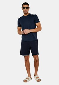 Marinblå t-shirt med korta ärmar, kombinerad med svarta ribbade shorts med vit kant, och beigea slidesandaler. Tillbehören inkluderar svarta solglasögon.